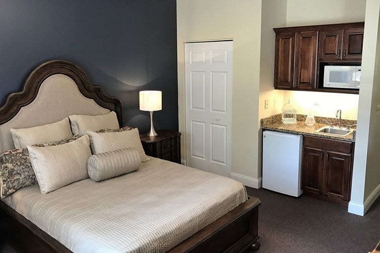 A cozy bedroom featuring a large bed with multiple pillows, a nightstand with a lamp, and a small kitchenette with a refrigerator.