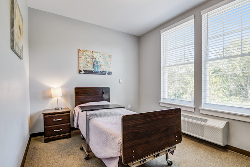 A well-lit patient room featuring a hospital bed, nightstand, and natural light from the window.