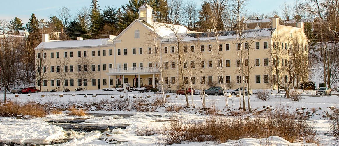 A large building surrounded by snow-covered ground and trees, with a frozen lake in the foreground.