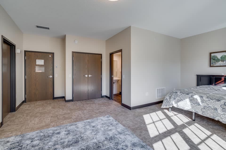 A spacious bedroom with carpet flooring, featuring doors and a window allowing natural light.