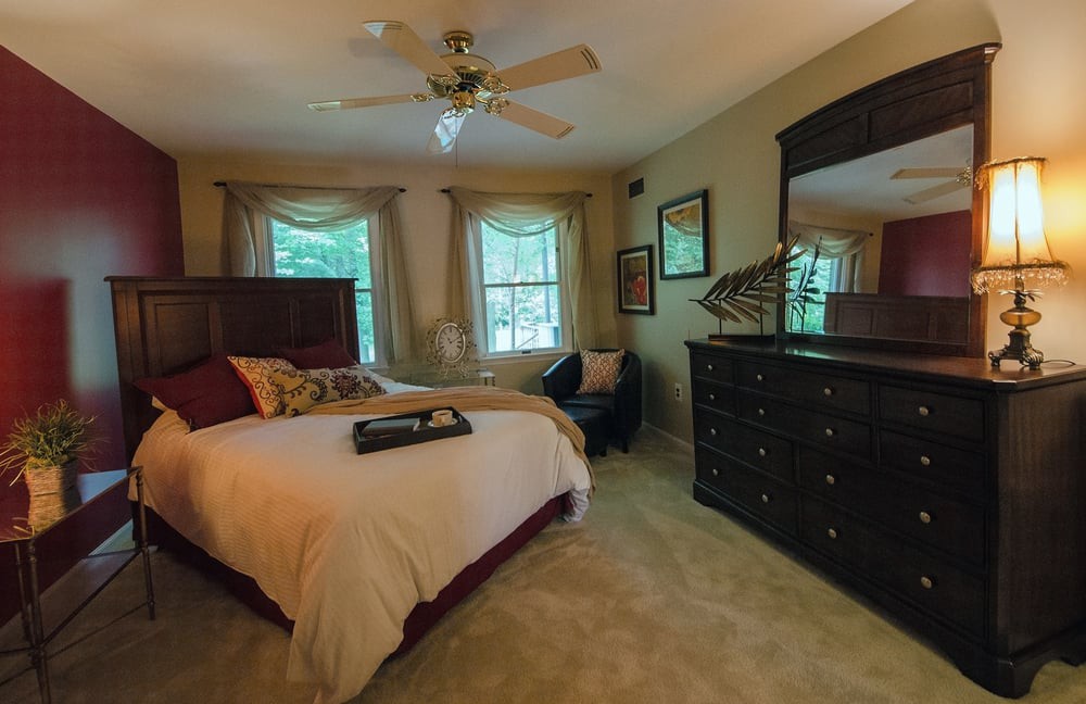 A cozy bedroom with elegant furniture, including a bed, dresser, and mirror, complemented by natural light from two windows.