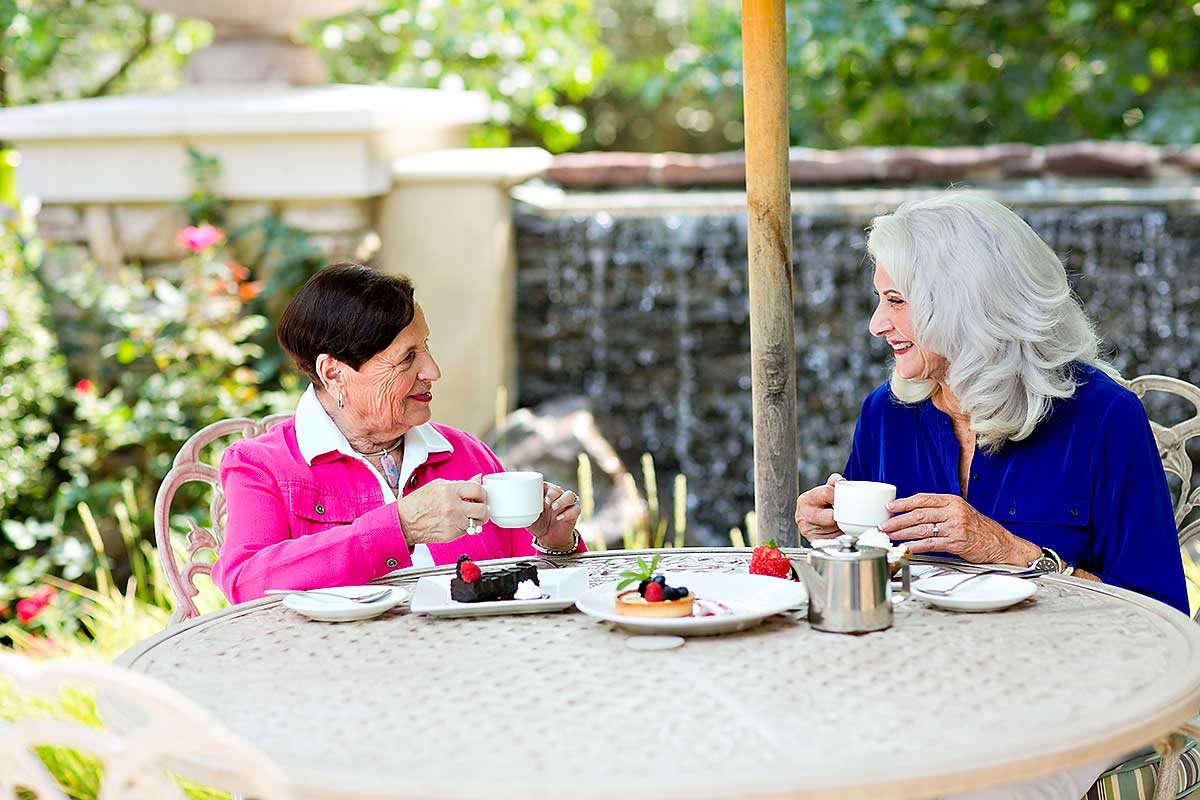 Two elderly women enjoying tea and desserts in a garden setting.