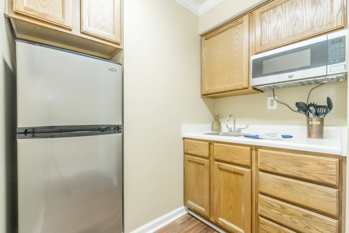 A compact apartment kitchen with wooden cabinets, a fridge, microwave, and sink.