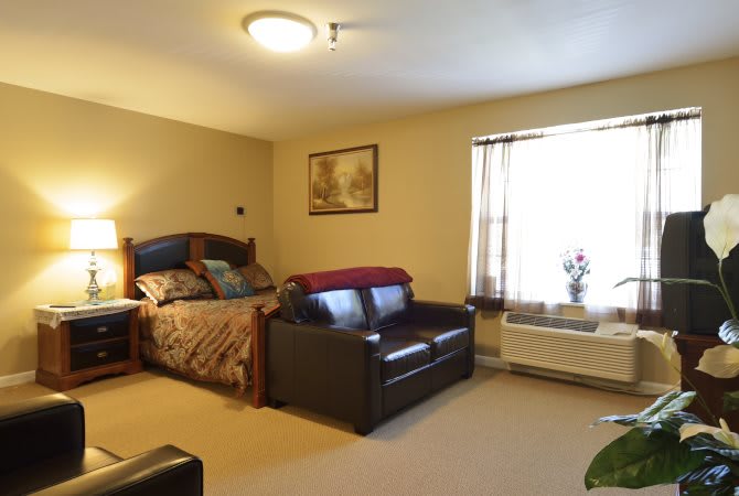 A cozy bedroom with a bed and sofa, featuring soft lighting and a window letting in natural light.