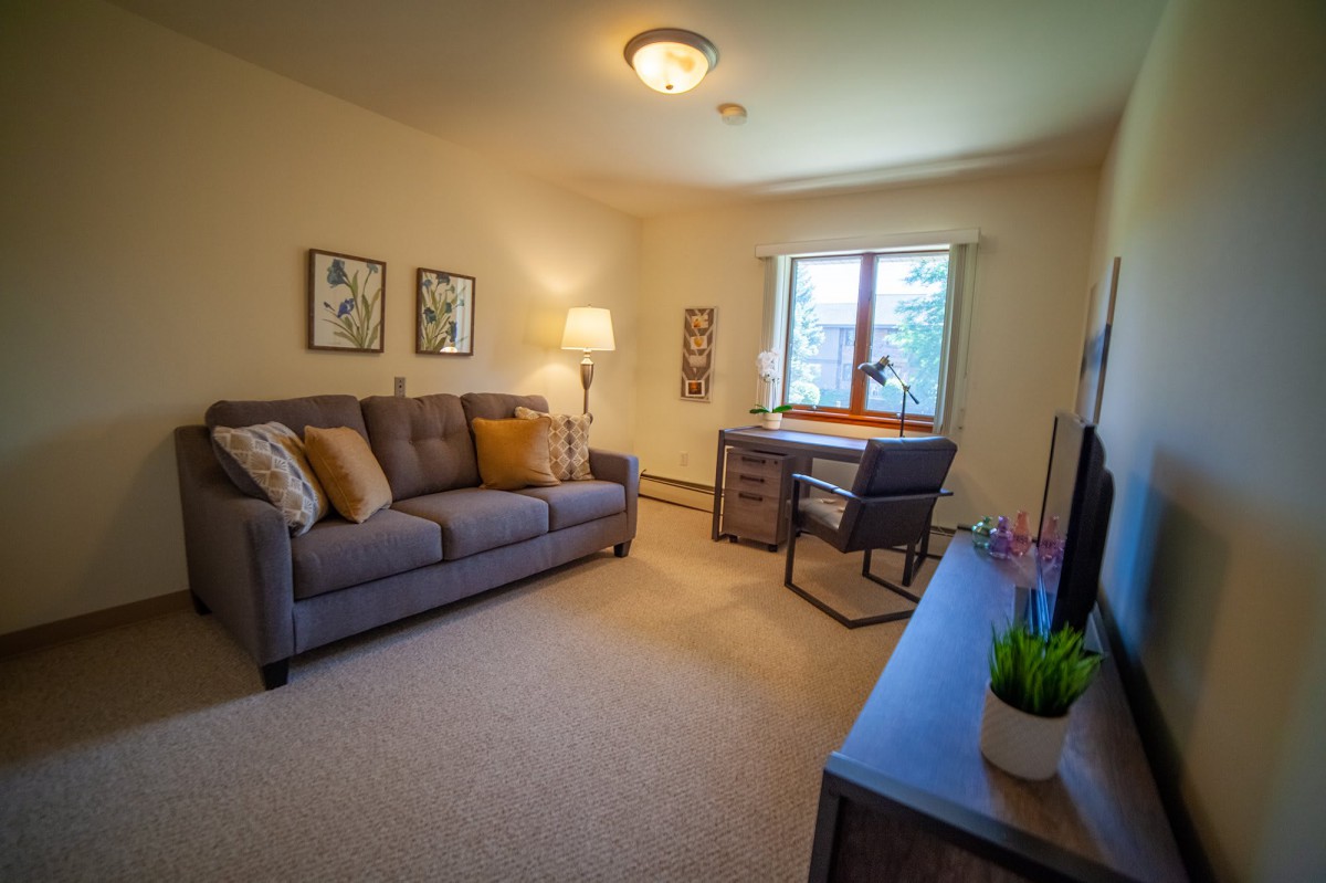 A cozy living room with a sofa, desk, and large window, featuring warm lighting and decor.