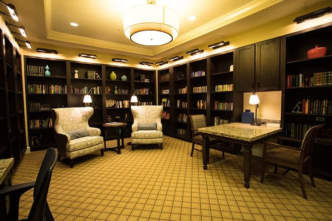 A cozy library room with bookshelves, armchairs, and warm lighting.