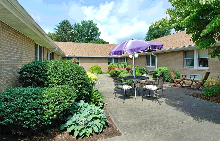 A tranquil patio area with outdoor furniture and a purple umbrella, surrounded by lush greenery.