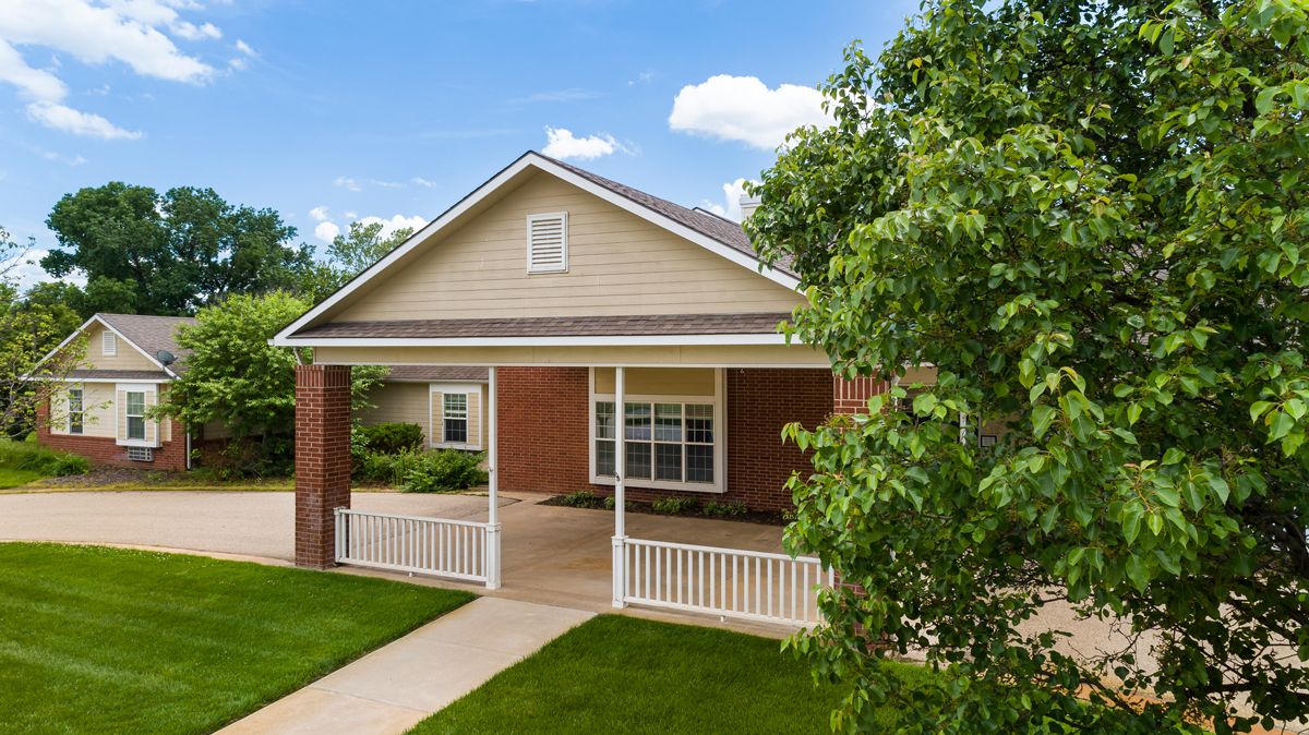 A charming suburban house with a brick exterior and a covered porch surrounded by lush greenery.