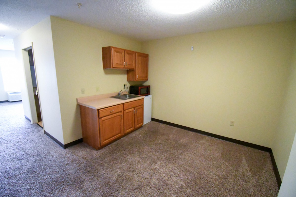 A small kitchenette with wooden cabinets, a sink, and carpeted flooring in a room with yellow walls.