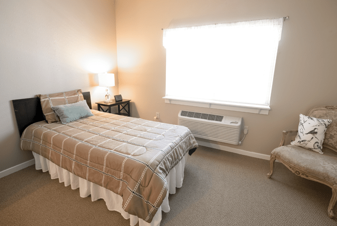 A cozy bedroom with a neatly made single bed, soft lighting from a lamp, and natural light coming through the window.
