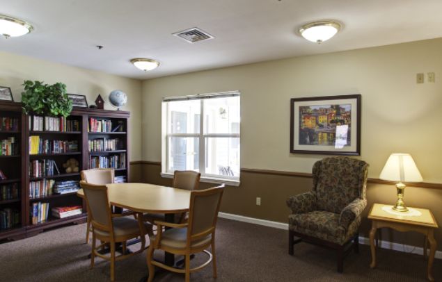 A cozy reading room with a table and chairs, featuring a bookshelf and window.