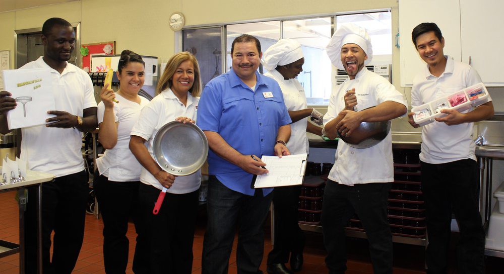 A group of chefs and a person holding a certificate, posing in a kitchen with cooking utensils.