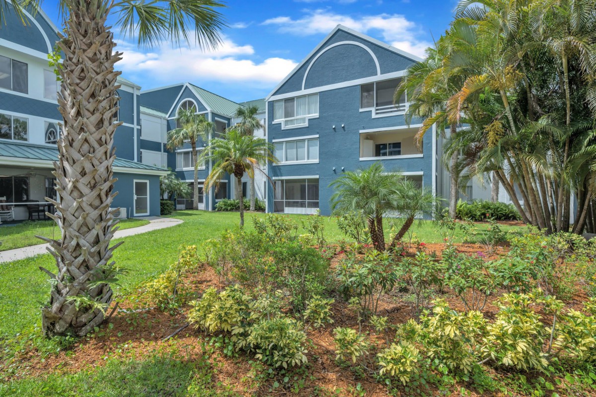 A scenic view of modern apartment buildings surrounded by lush greenery and palm trees under a bright, sunny sky.