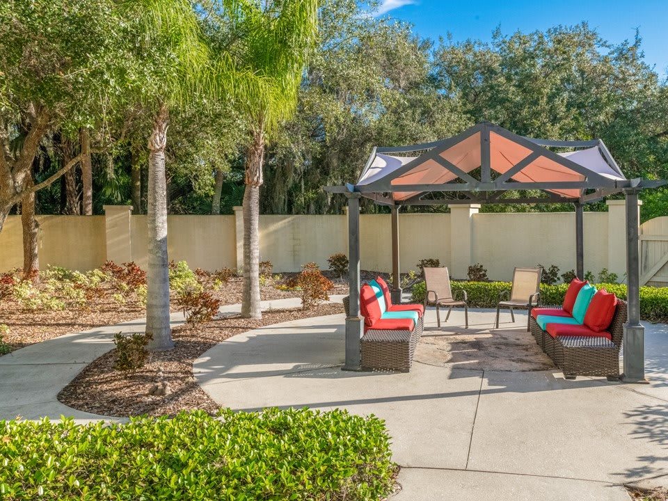 A sunny outdoor patio area with a gazebo, colorful seating, and lush landscaping.