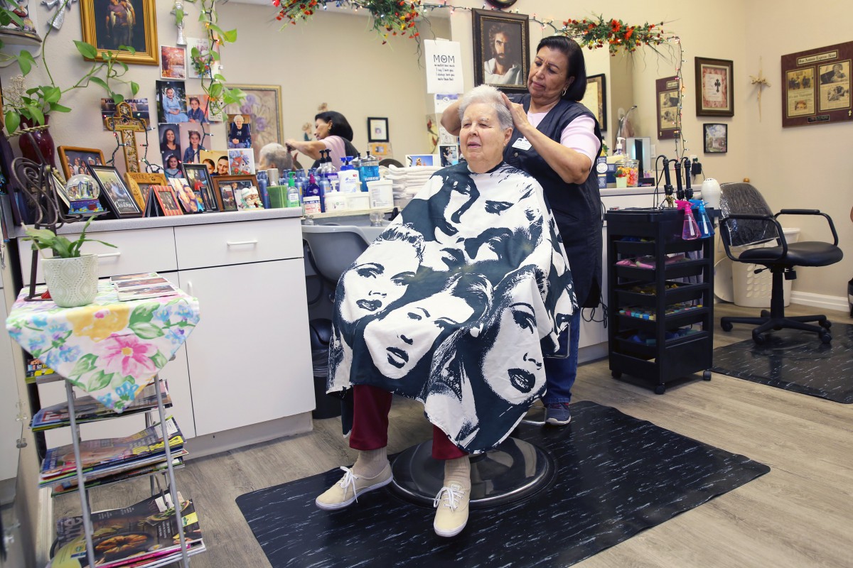 A hairstylist is working on a customer's hair in a cozy salon, with the customer wearing a cape featuring printed faces.