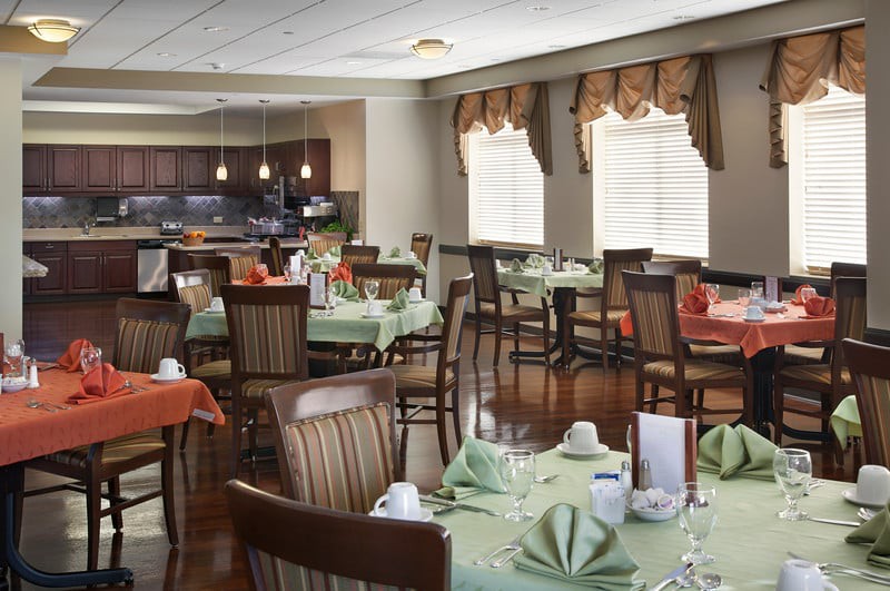 A well-lit dining room with neatly arranged tables and chairs, set with green napkins and crockery.