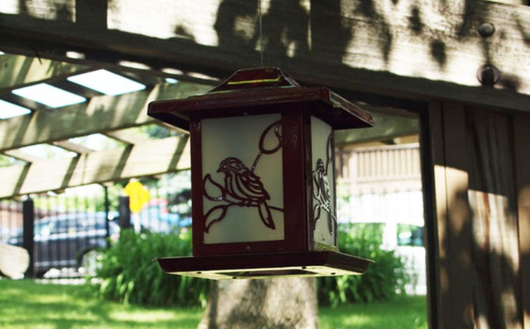 A decorative outdoor lantern with a bird design, hanging in a garden.