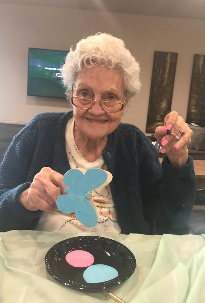An elderly woman smiles while decorating a blue butterfly-shaped cookie.
