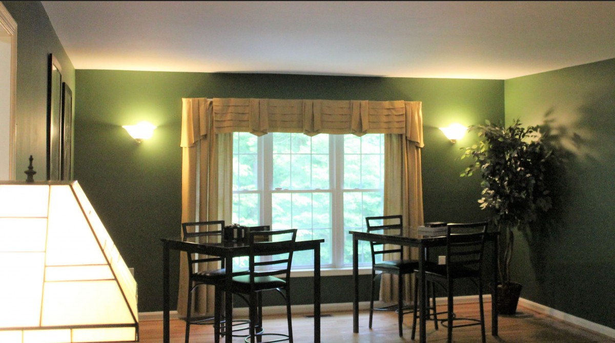 A cozy dining area with dark green walls, featuring multiple tables and chairs, illuminated by wall lamps and natural light from a large window with beige curtains.