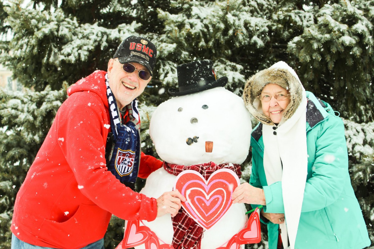A cheerful couple poses with a snowman holding a heart in a snowy setting.