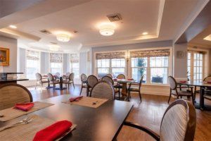 A bright dining room with multiple tables and chairs, featuring large windows allowing natural light to fill the space.