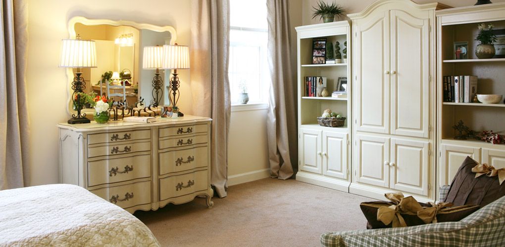 A cozy bedroom with traditional furniture, featuring a dresser with a lamp, and a wardrobe by the window.