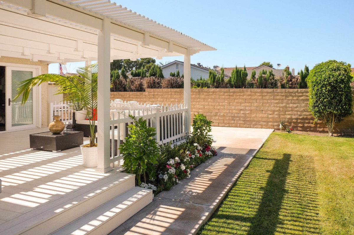A serene backyard patio with a white pergola, surrounded by lush greenery and a well-manicured lawn.