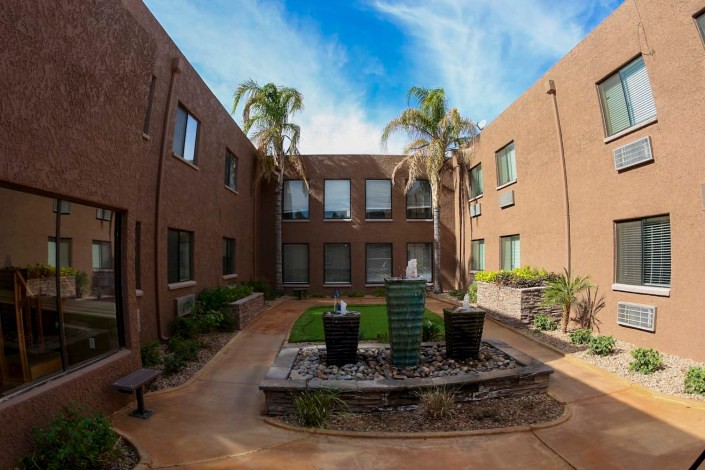 A serene courtyard with a central fountain, surrounded by palm trees and a two-story brick building.