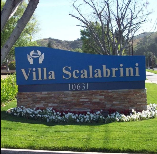 A blue entrance sign for Villa Scalabrini with well-maintained landscaping.
