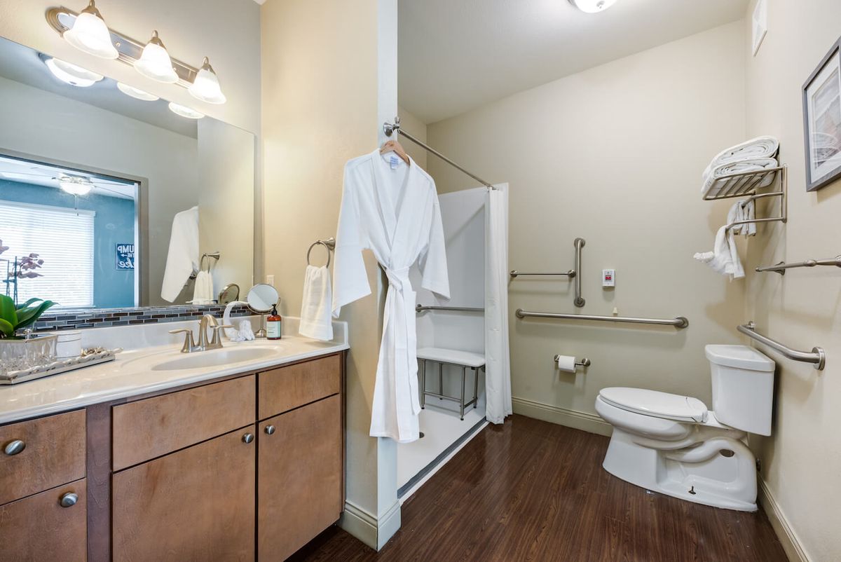 A modern bathroom with accessibility features, including a shower bench and grab bars, with a robe hanging near the sink.