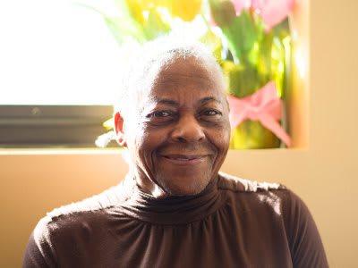 An elderly person smiling warmly indoors with soft natural light coming through the window.