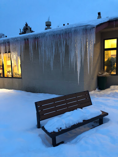 A serene winter scene featuring a bench covered in snow, icicles hanging from the roof, and a warm glow from the windows.