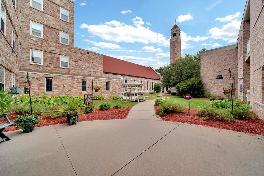 A courtyard with a pathway leading to buildings and a tall tower under a blue sky with clouds.