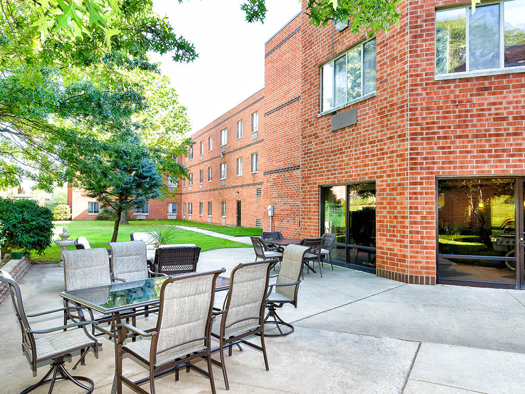 A serene outdoor patio area with chairs and tables beside a brick building, surrounded by lush greenery.