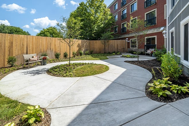 A beautifully landscaped garden with a circular paved path, surrounded by greenery and bordered by a wooden fence, adjacent to a building.