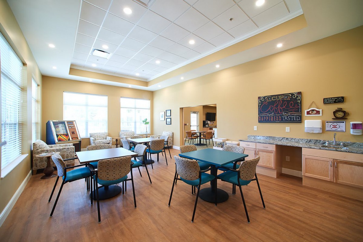 A spacious, well-lit room with multiple tables and chairs, featuring large windows and a kitchenette.