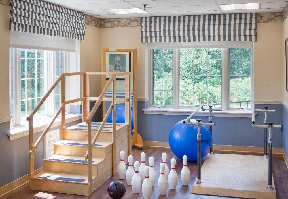 A physical therapy room with exercise equipment including stairs, a stability ball, parallel bars, and bowling pins near large windows.
