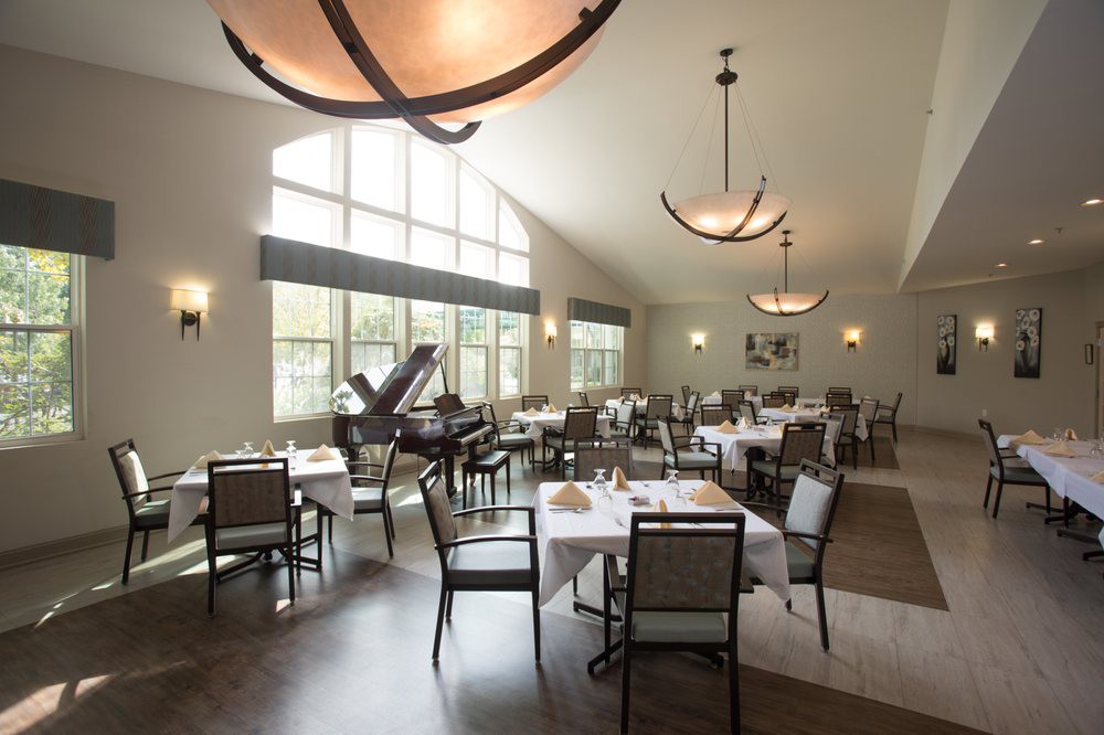 A spacious and elegantly designed restaurant interior with neatly arranged tables and chairs, large windows, and a grand piano in the corner.