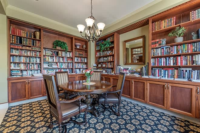 A cozy library with wooden bookshelves, a round table, and upholstered chairs under a chandelier.