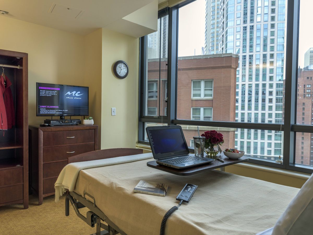 A hospital room featuring a patient bed with a laptop and flowers, overlooking an urban cityscape through large windows.