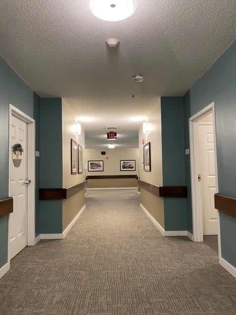 A well-lit hallway with carpeted flooring, featuring doors on either side and framed pictures on the walls.