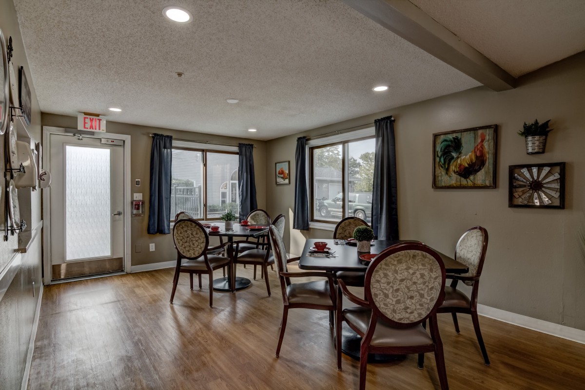 A cozy dining area with tables and chairs, featuring large windows and wooden flooring.