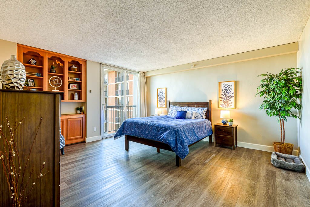 A cozy bedroom with a wooden floor, featuring a bed with blue bedding and nightstands on each side, illuminated by warm lighting.