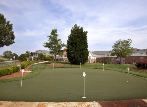 A well-maintained putting green in a residential area with houses and trees in the background.