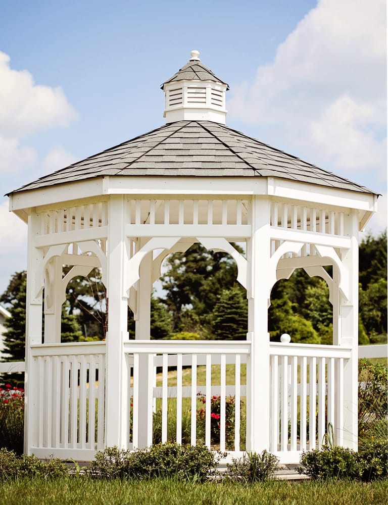 A white gazebo set in a garden with lush greenery and a clear sky.