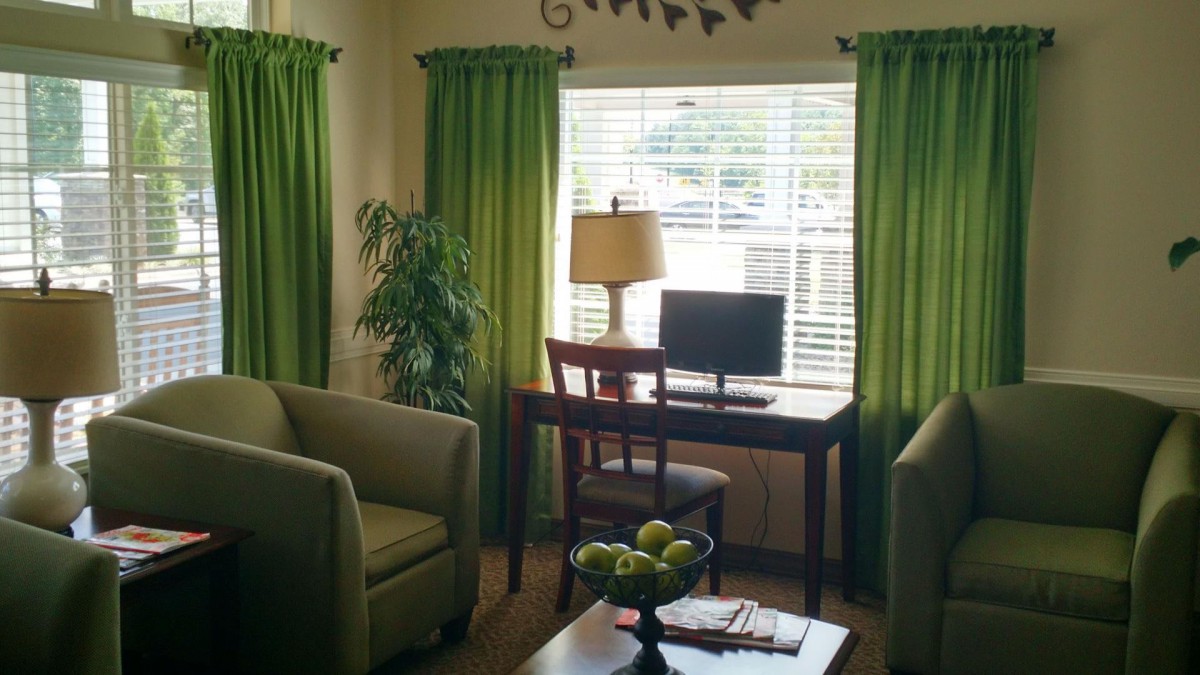 A cozy living room with a green color theme, featuring a desk with a computer and lamp by the window.