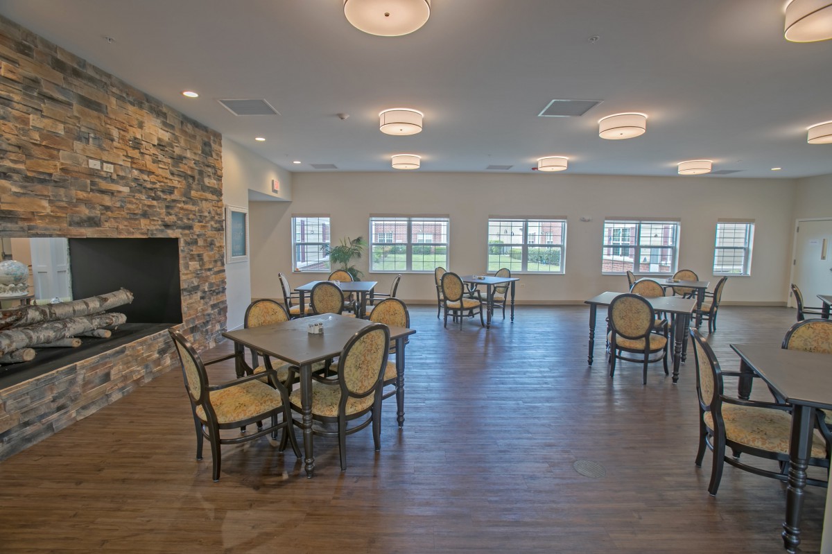 A spacious dining room with several tables and chairs, featuring large windows, wood flooring, and a stone fireplace.