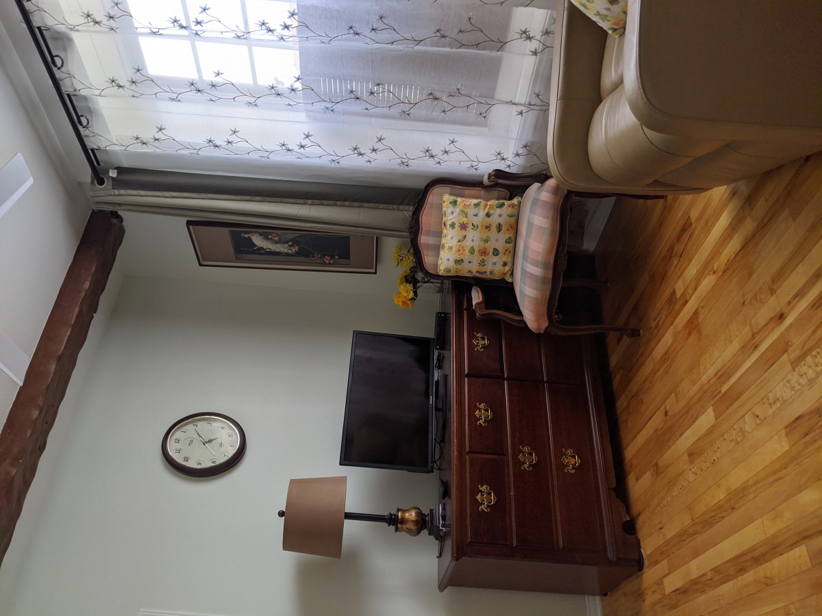 A cozy living room featuring a TV on a wooden dresser, a plaid upholstered chair, a lamp, and decorative curtains.