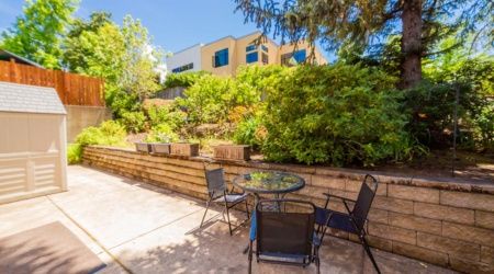 A sunny patio area with a glass table and four chairs, surrounded by lush greenery.