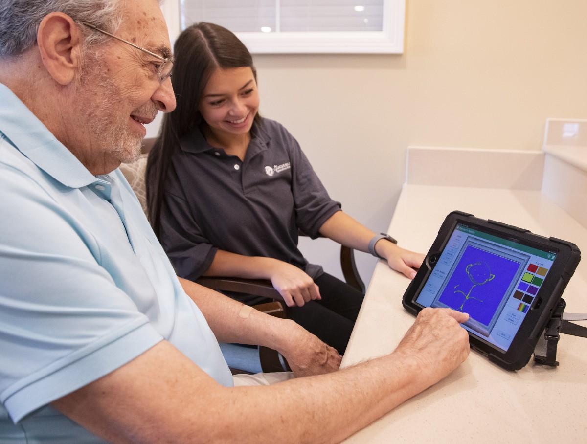 An elderly man and a young woman interact with a tablet, focusing on learning new technology.